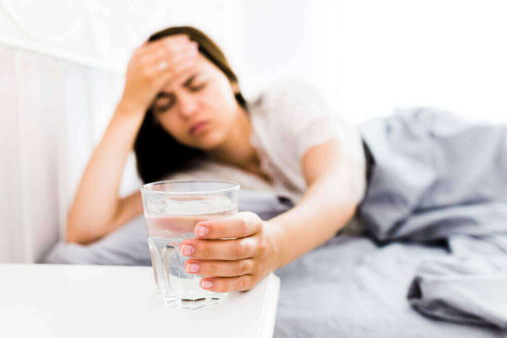 숙취 때문에 머리를 부여잡고 있는 여자의 모습. 술 빨리 깨는 방법 중 물을 마시기 위해 물이 든 컵을 잡고 있다.
